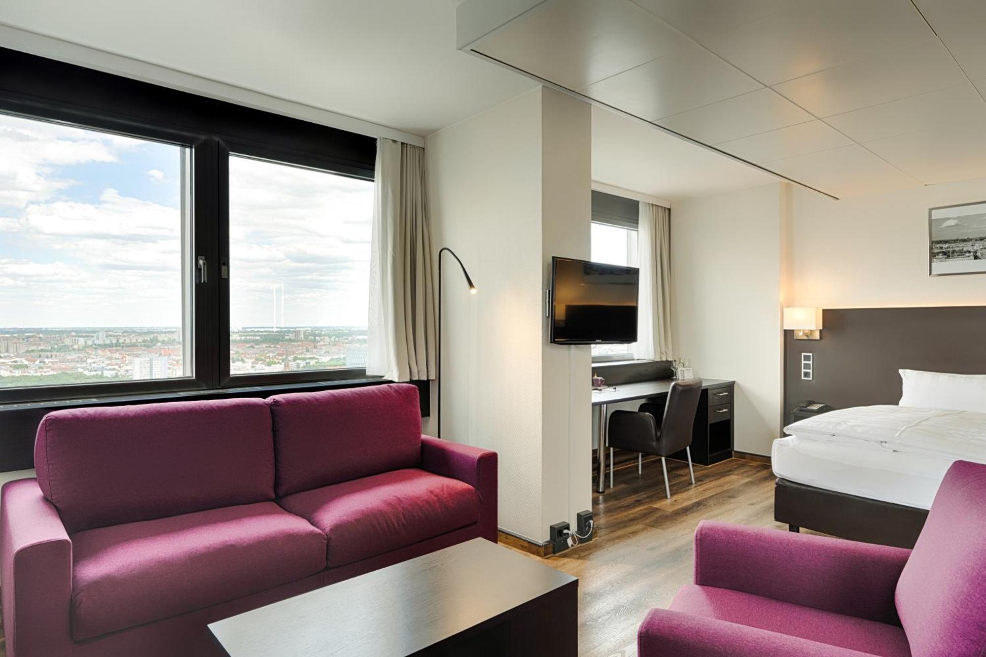 Park Inn By Radisson Berlin Alexanderplatz ภายนอก รูปภาพ The photo shows a modern hotel room with a stylish design. It features a seating area with two purple sofas and a coffee table. Opposite the seating area, there is a desk with a chair and a television mounted on the wall. The room has large windows a