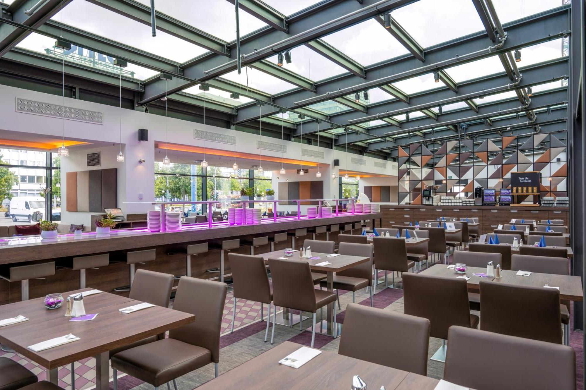 Park Inn By Radisson Berlin Alexanderplatz ภายนอก รูปภาพ The photo shows the interior of a modern restaurant. It features a spacious and bright layout with large windows allowing natural light to flow in. The ceiling is made of glass panels, enhancing the open atmosphere. 

The dining area has several dark