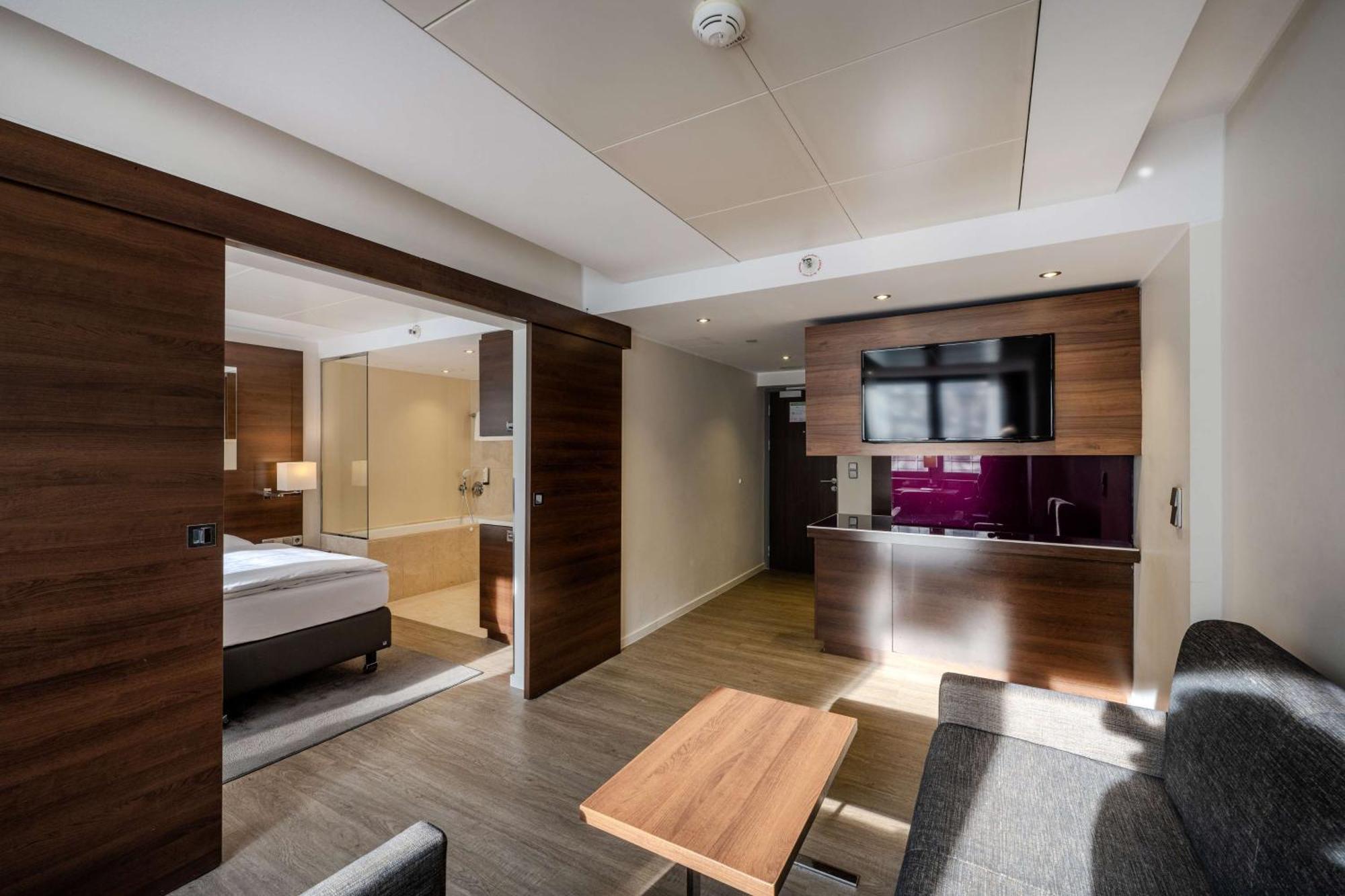 Park Inn By Radisson Berlin Alexanderplatz ภายนอก รูปภาพ The photo shows a modern hotel room with a well-designed layout. On one side, there is a bedroom area featuring a bed and light-colored bedding. The room is separated by sliding wooden doors. The living area includes a comfortable gray couch and a wo