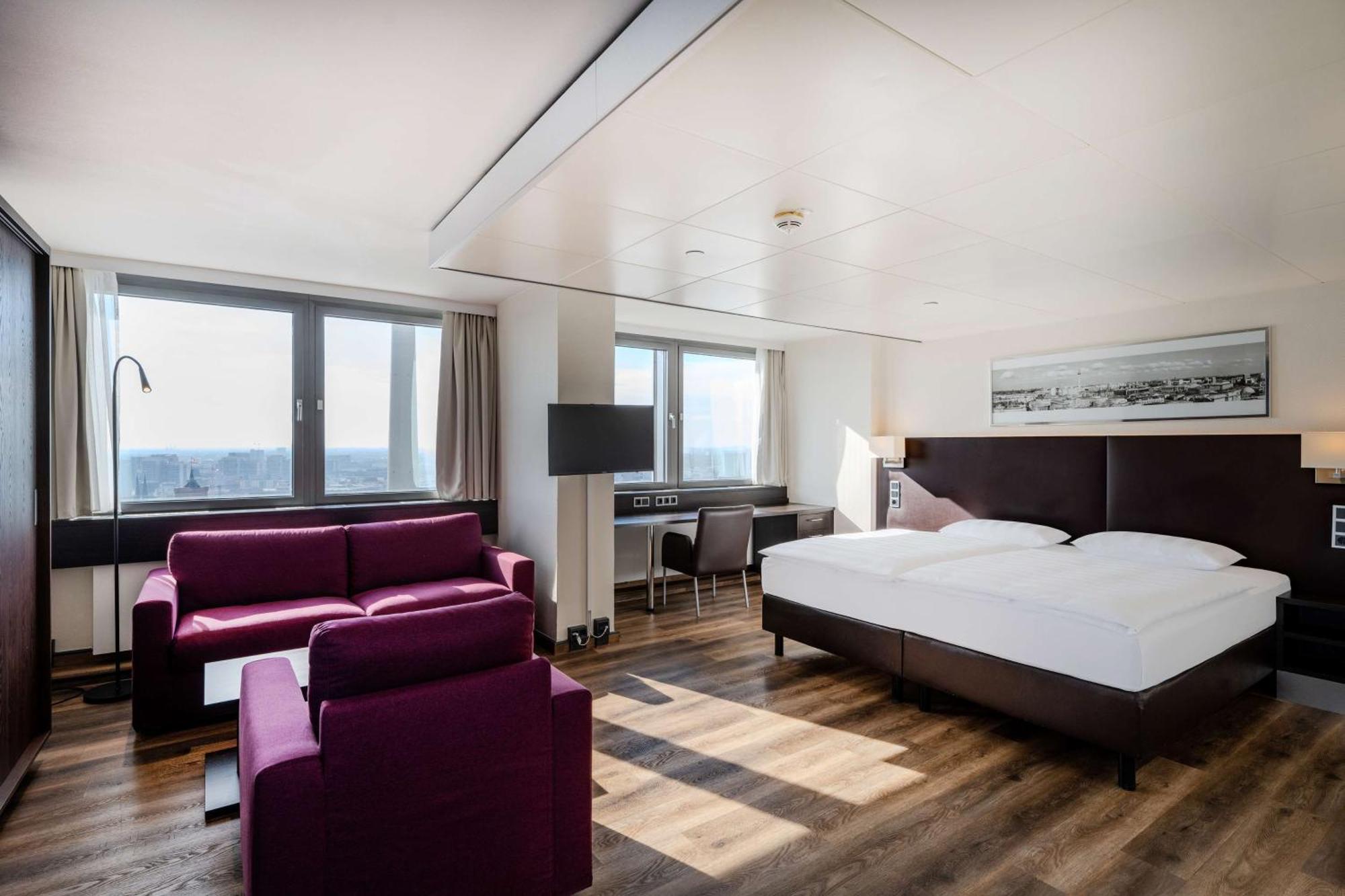Park Inn By Radisson Berlin Alexanderplatz ภายนอก รูปภาพ The photo shows a modern hotel room featuring a large bed with white bedding. There's a cozy seating area with a purple sofa and armchair. A desk and chair are positioned near a window, which provides natural light and offers a view of the outside. T