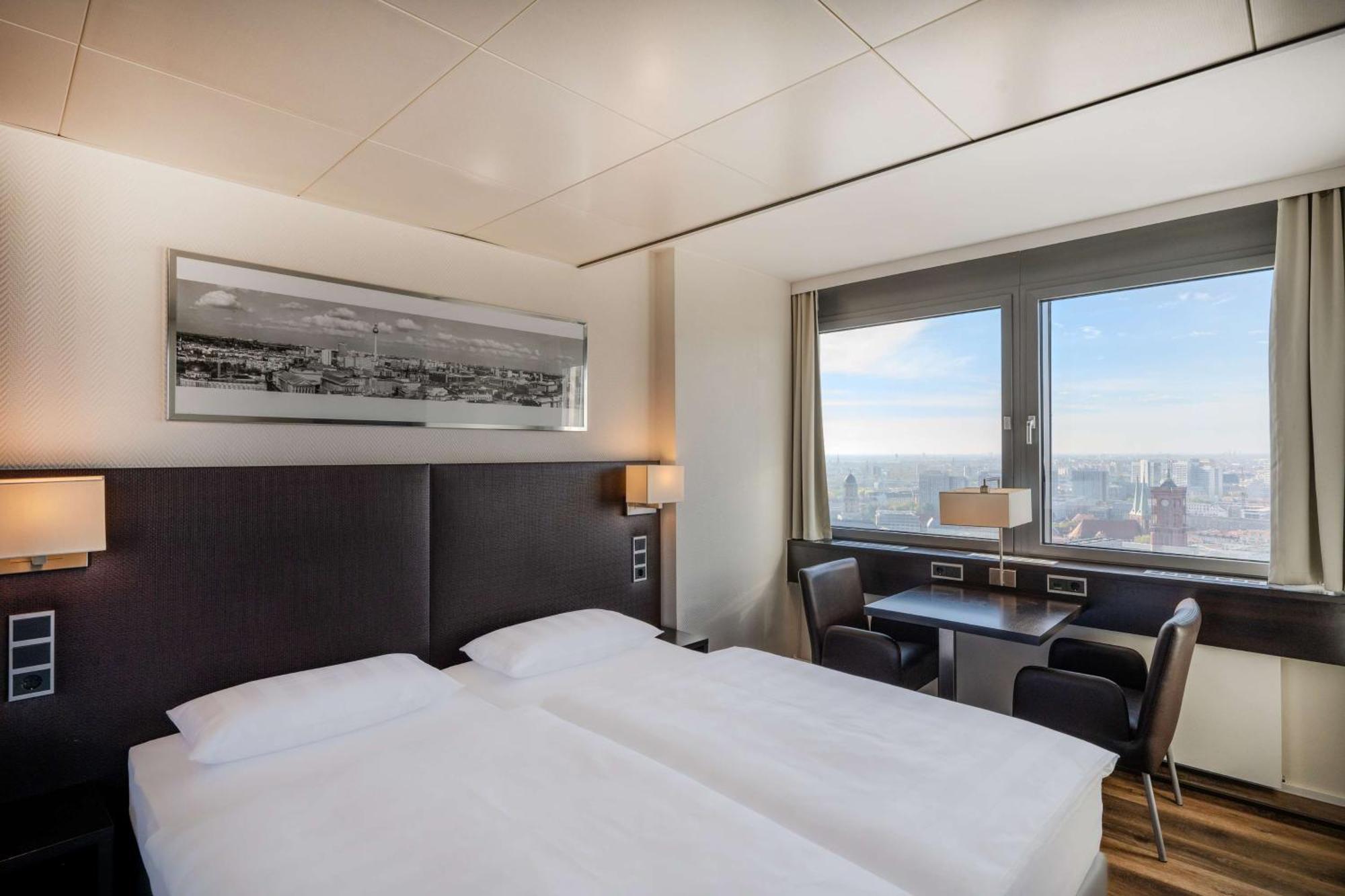 Park Inn By Radisson Berlin Alexanderplatz ภายนอก รูปภาพ The photo shows a modern hotel room featuring two single beds with white linens and dark headboards. There is a large window that provides natural light and a view of the cityscape outside. The room is equipped with a small table and two black chairs